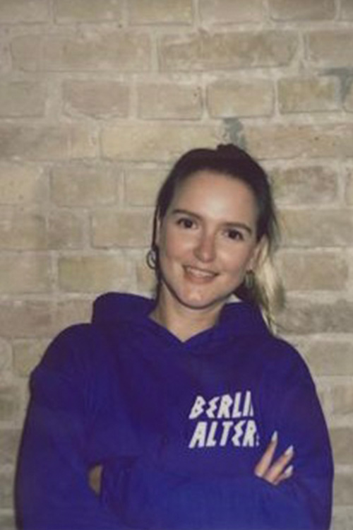 Frau lächelt in die Kamera, trägt blauen Kapuzenpullover mit Aufdruck "BERLIN ALTER", vor gemauerter Wand.