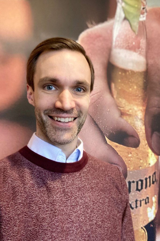 Mann mit braunem Pulli lächelt vor einem Werbeplakat mit einer Corona-Bierflasche.