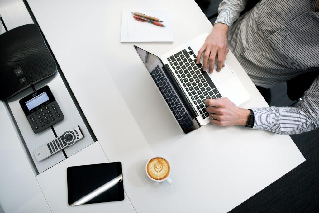 Mann tippt auf Laptop in Büroumgebung mit Tablet, Telefon, Konferenztelefon und Kaffeetasse.